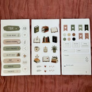 Könnte beinhalten: Drei Stickerbögen für Bücherliebhaber. Die Aufkleber zeigen Buchgrafiken, darunter Bücherstapel, offene Bücher und Kaffeetassen. Die Bögen enthalten auch Etiketten wie "TBR", "Currently Reading" und "Favorite."