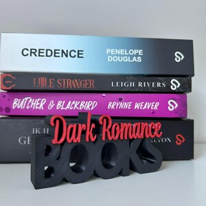 May include: A stack of four books with titles including "Credence" and "Little Stranger." A black and red "Dark Romance Books" sign is in front of the books. The books have a variety of colors, including blue, black, and purple.