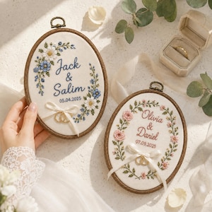 May include: Two oval wooden embroidery hoops featuring white fabric with floral embroidery, names, and dates. One hoop displays Jack & Salim 05.04.2025 with blue flowers, the other Olivia & Daniel 07.09.2025 with pink flowers. Both include white ribbons holding gold rings.