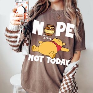 Puede incluir: Una camiseta marrón con un gráfico blanco de Winnie the Pooh durmiendo con un tarro de miel que dice "Hunny". El texto "NOPE" está por encima del oso y "NOT TODAY" está por debajo del oso.