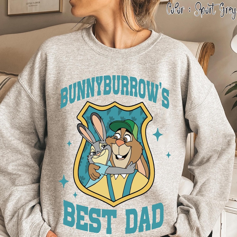 Puede incluir: Sudadera gris deportiva con un gr&aacute;fico de dibujos animados de dos conejos abraz&aacute;ndose, con el texto "BUNNYBURROW'S BEST DAD" en color turquesa. El gr&aacute;fico est&aacute; dentro de un escudo. La sudadera es de color gris claro.