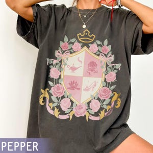 Puede incluir: Camiseta gris oscuro con un dise&ntilde;o de escudo con una corona, rosas rosas y un escudo rosa y blanco. El escudo tiene cuatro cuadrantes con ilustraciones de una l&aacute;mpara, una concha, una flor de loto y un zapato de cristal. La palabra "PEPPER" est&aacute; en la parte inferior.