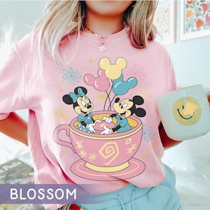 May include: Pale pink t-shirt with a graphic of Mickey and Minnie Mouse in a teacup with Mickey Mouse balloons. The shirt has the word "BLOSSOM" at the bottom. The t-shirt is a light pink colour.