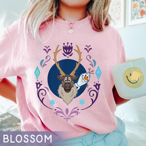 Puede incluir: Camiseta rosa claro con un dise&ntilde;o de Sven y Olaf de la pel&iacute;cula Frozen, enmarcado por elementos decorativos. La palabra "BLOSSOM" est&aacute; impresa en la parte inferior. La camiseta es de estilo casual.