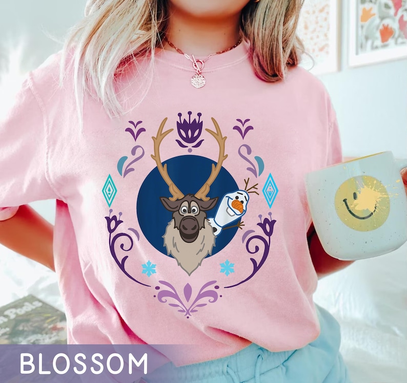 Puede incluir: Camiseta rosa claro con un dise&ntilde;o de Sven y Olaf de la pel&iacute;cula Frozen, enmarcado por elementos decorativos. La palabra "BLOSSOM" est&aacute; impresa en la parte inferior. La camiseta es de estilo casual.