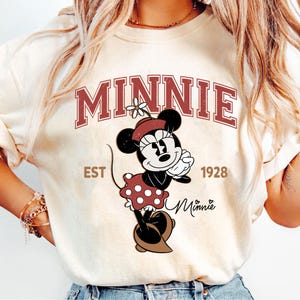 May include: Cream-colored sweatshirt featuring a graphic of Minnie Mouse in a red polka-dot dress and a matching hat. The word "MINNIE" is arched across the top in maroon letters, with "EST 1928" below. The Minnie Mouse graphic is in the center.