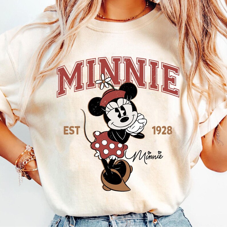 May include: Cream-colored sweatshirt featuring a graphic of Minnie Mouse in a red polka-dot dress and a matching hat. The word "MINNIE" is arched across the top in maroon letters, with "EST 1928" below. The Minnie Mouse graphic is in the center.