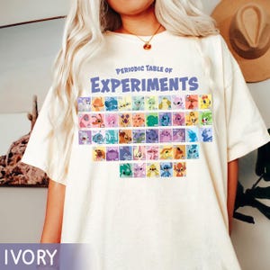 Könnte beinhalten: Elfenbeinfarbenes T-Shirt mit dem blauen Text "Periodic Table of Experiments". Das Shirt zeigt eine farbenfrohe Grafik im Periodensystem-Stil mit Zeichentrickfiguren. Das Shirt hat eine lockere Passform.