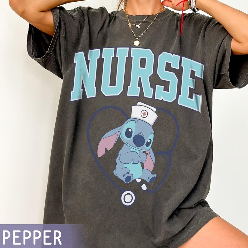 May include: Dark gray t-shirt with the word "NURSE" in light blue block letters. Below the text is a cartoon character wearing a nurse's hat, inside a blue heart with a stethoscope. The shirt is a casual style.