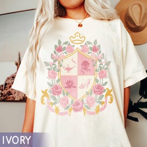 Puede incluir: Camiseta de color marfil con un dise&ntilde;o de escudo con una corona, rosas y cintas. El escudo est&aacute; dividido en cuatro secciones, cada una con una ilustraci&oacute;n diferente en rosa y dorado. La palabra "IVORY" est&aacute; impresa en la parte inferior.