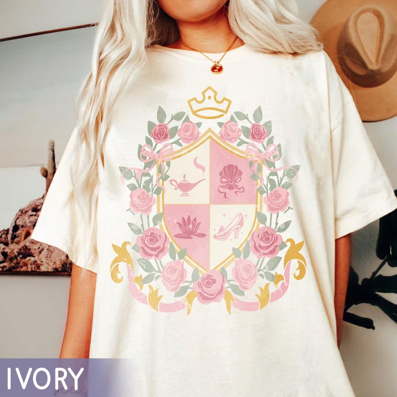Puede incluir: Camiseta de color marfil con un dise&ntilde;o de escudo con una corona, rosas y cintas. El escudo est&aacute; dividido en cuatro secciones, cada una con una ilustraci&oacute;n diferente en rosa y dorado. La palabra "IVORY" est&aacute; impresa en la parte inferior.