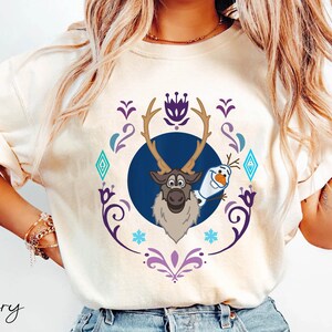 Puede incluir: Camiseta de color marfil con un dise&ntilde;o de dibujos animados de Sven el reno y Olaf el mu&ntilde;eco de nieve de la pel&iacute;cula Frozen. El dise&ntilde;o est&aacute; enmarcado por un borde decorativo p&uacute;rpura y azul. La palabra "Ivory" est&aacute; impresa en la parte inferior.