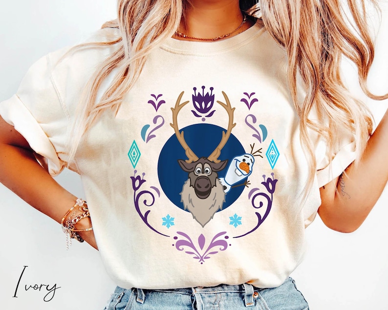 Puede incluir: Camiseta de color marfil con un dise&ntilde;o de dibujos animados de Sven el reno y Olaf el mu&ntilde;eco de nieve de la pel&iacute;cula Frozen. El dise&ntilde;o est&aacute; enmarcado por un borde decorativo p&uacute;rpura y azul. La palabra "Ivory" est&aacute; impresa en la parte inferior.