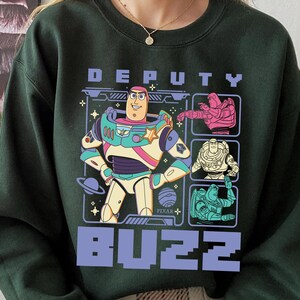 May include: Dark green sweatshirt featuring a graphic of Buzz Lightyear from Toy Story. The design includes the word "DEPUTY" above and "BUZZ" below the character. The Buzz Lightyear graphic is in a colorful, cartoon style.