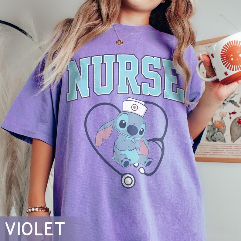 May include: Violet t-shirt with the word "NURSE" in light blue block letters. Below the text is a cartoon character wearing a nurse's hat, inside a stethoscope-shaped heart. The word "VIOLET" is printed at the bottom of the shirt.