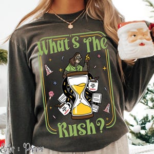 Puede incluir: Camiseta de manga larga color pimienta con un gr&aacute;fico que presenta un perezoso, un reloj de arena y el texto "What's The Rush?". El dise&ntilde;o incluye tazas de caf&eacute; y elementos de carretera. La camiseta tambi&eacute;n tiene el texto "You want it when?"