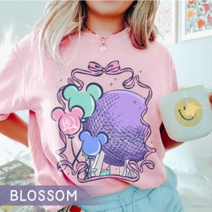 May include: Pink t-shirt with a graphic of a purple sphere, Mickey Mouse-shaped balloons in blue, green, and pink, and the text "BLOSSOM". A white mug with a yellow smiley face is held in the hand.