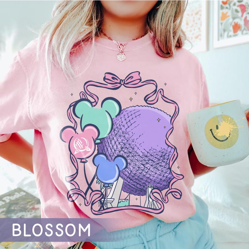 May include: Pink t-shirt with a graphic of a purple sphere, Mickey Mouse-shaped balloons in blue, green, and pink, and the text "BLOSSOM". A white mug with a yellow smiley face is held in the hand.