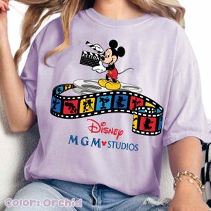 Pu&ograve; includere: T-shirt color orchidea con Topolino che tiene una clapperboard. Il design include una bobina di film e una striscia di film colorata con personaggi Disney. Il testo "Disney MGM Studios" &egrave; stampato sotto.