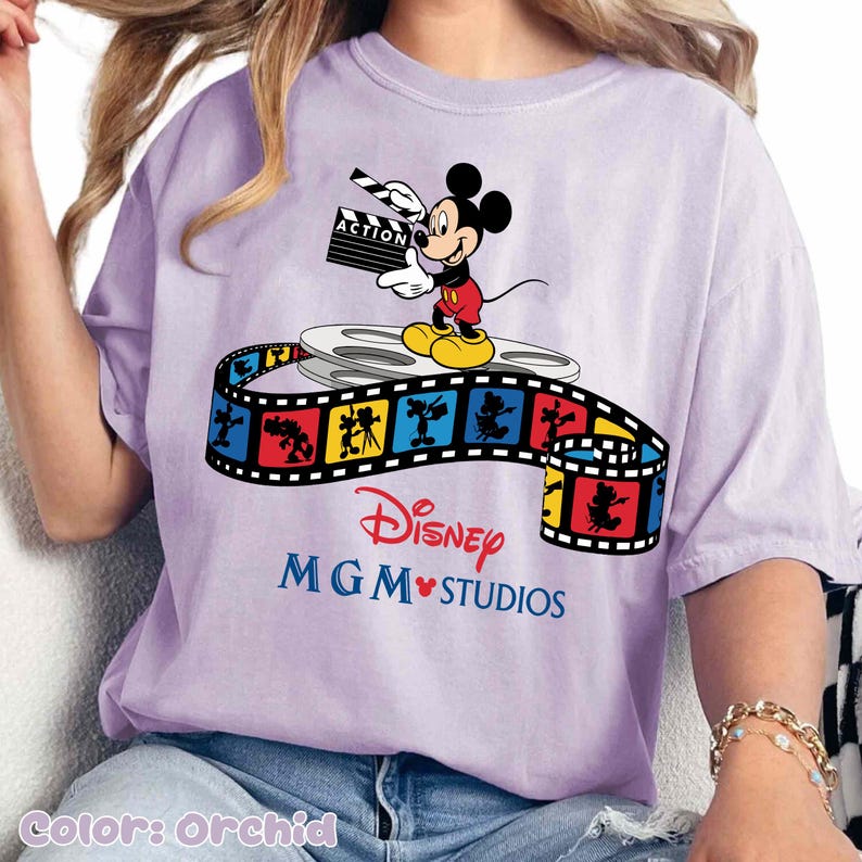 Pu&ograve; includere: T-shirt color orchidea con Topolino che tiene una clapperboard. Il design include una bobina di film e una striscia di film colorata con personaggi Disney. Il testo "Disney MGM Studios" &egrave; stampato sotto.