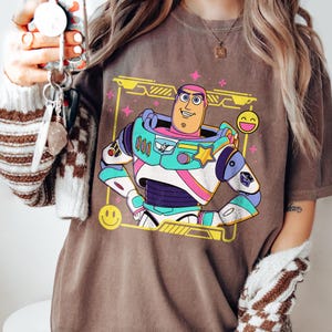 May include: Brown t-shirt featuring a colorful graphic of Buzz Lightyear from Toy Story. The design includes a yellow frame, pink stars, and a smiling emoji. The shirt is a comfortable, casual wear item.