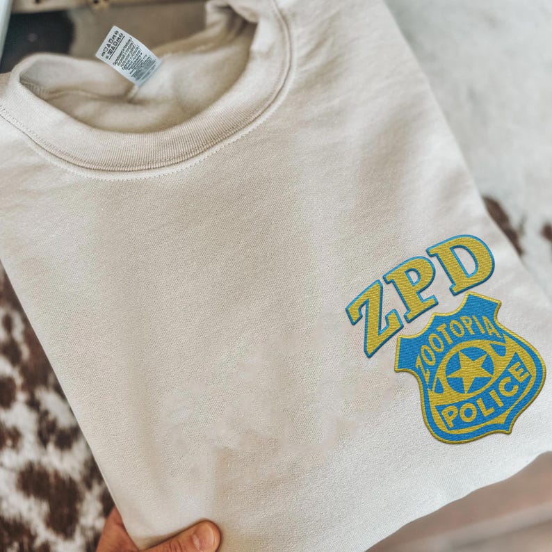 May include: A beige crewneck sweatshirt featuring a blue and yellow embroidered ZPD Zootopia Police badge graphic on the lower right side. The garment has a ribbed collar and a white fabric tag attached to the inner neckline with visible text.
