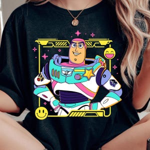 May include: Black t-shirt featuring a colorful cartoon illustration of Buzz Lightyear from Toy Story. The design includes a yellow frame, pink accents, and a smiling face. The character is depicted in his iconic space ranger suit.