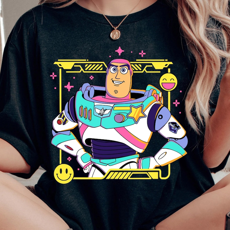 May include: Black t-shirt featuring a colorful cartoon illustration of Buzz Lightyear from Toy Story. The design includes a yellow frame, pink accents, and a smiling face. The character is depicted in his iconic space ranger suit.