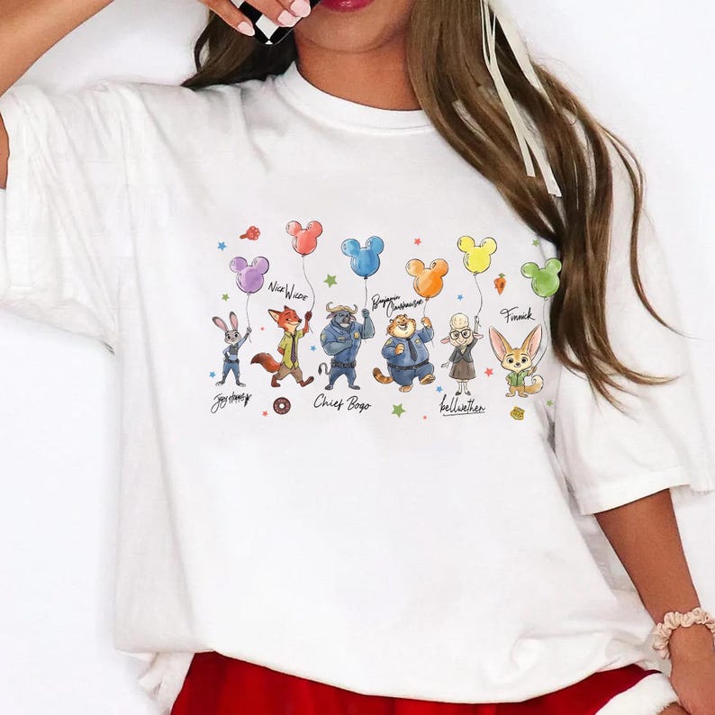 Op de afbeelding: Wit T-shirt met een kleurrijke afbeelding van stripfiguren die ballonvormige ballonnen vasthouden. De personages zijn onder meer een konijn, een vos, een stier, een tijger, een schaap en een vos. De tekst bevat namen van personages en ballonvormen.