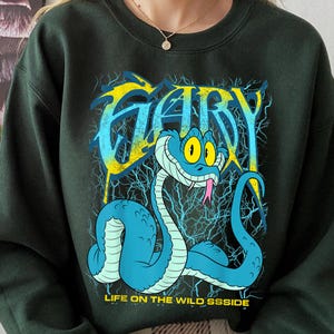 Op de afbeelding: Donkergroene sweatshirt met een cartoon slang ontwerp. Het ontwerp bevat de naam "GARY" in geel en blauw, en de tekst "LIFE ON THE WILD SSSSIDE". De slang is blauw met gele ogen en een roze tong.
