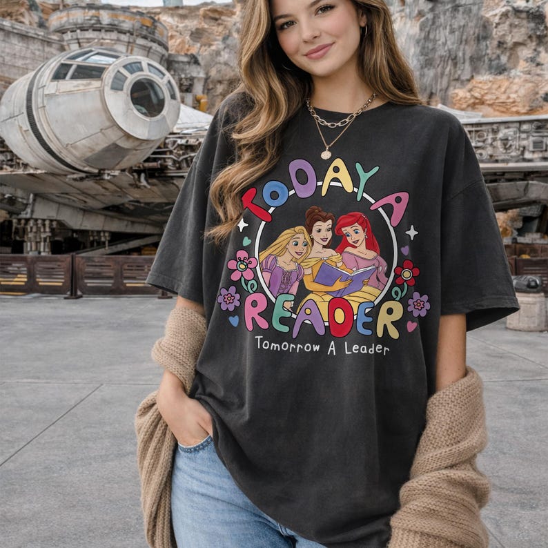 May include: Dark gray t-shirt with a colorful graphic that reads "Today a Reader, Tomorrow a Leader." The graphic features three cartoon princesses reading a book, surrounded by flowers. The shirt is oversized.