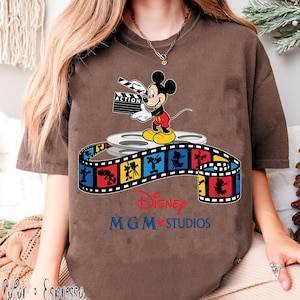 Pu&ograve; includere: T-shirt color espresso con Topolino che tiene una clapperboard con la scritta "ACTION". Mickey &egrave; in piedi su una bobina di film sopra una striscia di film colorata con personaggi Disney. La maglietta mostra anche "Disney MGM Studios".