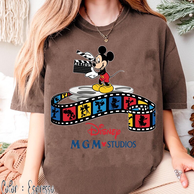 Pu&ograve; includere: T-shirt color espresso con Topolino che tiene una clapperboard con la scritta "ACTION". Mickey &egrave; in piedi su una bobina di film sopra una striscia di film colorata con personaggi Disney. La maglietta mostra anche "Disney MGM Studios".