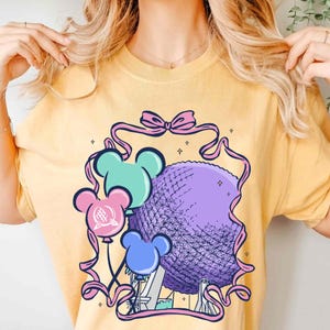 May include: Yellow t-shirt featuring a graphic of a purple sphere, resembling a geodesic dome, with Mickey Mouse-shaped balloons in teal, pink, and blue. The design is framed by a pink ribbon and bow, with small star accents.