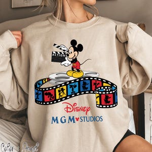 Pu&ograve; includere: Felpa beige con una grafica colorata di Topolino che tiene una clapperboard cinematografica. Il design include una bobina di film e una striscia di film con personaggi Disney. Il testo "Disney MGM Studios" &egrave; stampato sotto.
