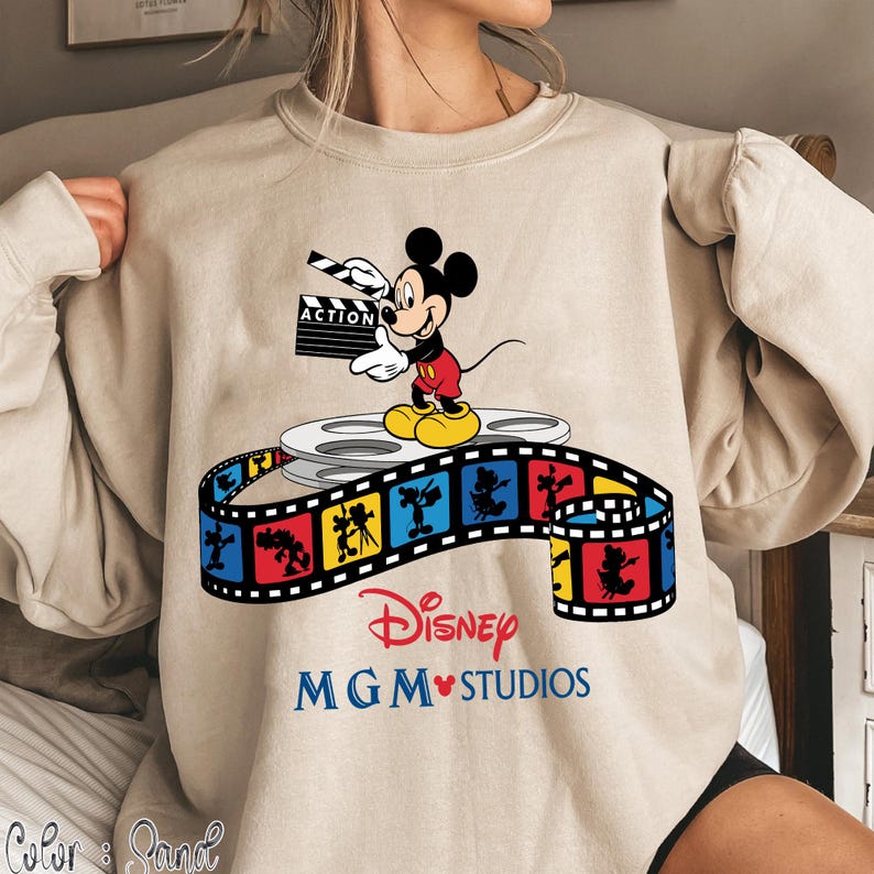 Pu&ograve; includere: Felpa beige con una grafica colorata di Topolino che tiene una clapperboard cinematografica. Il design include una bobina di film e una striscia di film con personaggi Disney. Il testo "Disney MGM Studios" &egrave; stampato sotto.