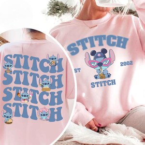 May include: A light pink sweatshirt featuring the word "STITCH" in blue block letters. The front of the sweatshirt has a graphic of the character Stitch wearing sunglasses and drinking a beverage. The back of the sweatshirt has multiple Stitch graphics.