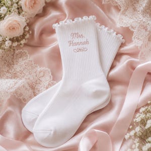 May include: A pair of white ribbed crew socks featuring a ruffled top edge. The socks are embroidered with the text Mrs. Hannah in a light pink script font, accompanied by a small decorative flourish design centered below the name.