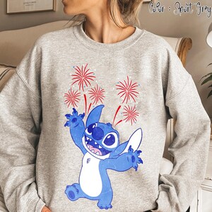 May include: Sport grey sweatshirt featuring a blue cartoon character with a big smile and outstretched arms, with red and white fireworks above. The text "Color: Sport Grey" is at the top right.