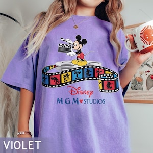 Pu&ograve; includere: Maglietta viola con una grafica di Topolino che tiene una clapperboard, in piedi su una bobina di film. Una striscia di film colorata con personaggi Disney avvolge la bobina. Il testo "Disney MGM Studios" &egrave; in basso.