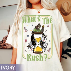 Puede incluir: Camiseta color marfil con un dise&ntilde;o gr&aacute;fico que presenta un perezoso se&ntilde;alando un reloj de arena. El texto "What's The Rush?" es verde, con gr&aacute;ficos adicionales de tazas de caf&eacute;, conos de tr&aacute;fico y flores.