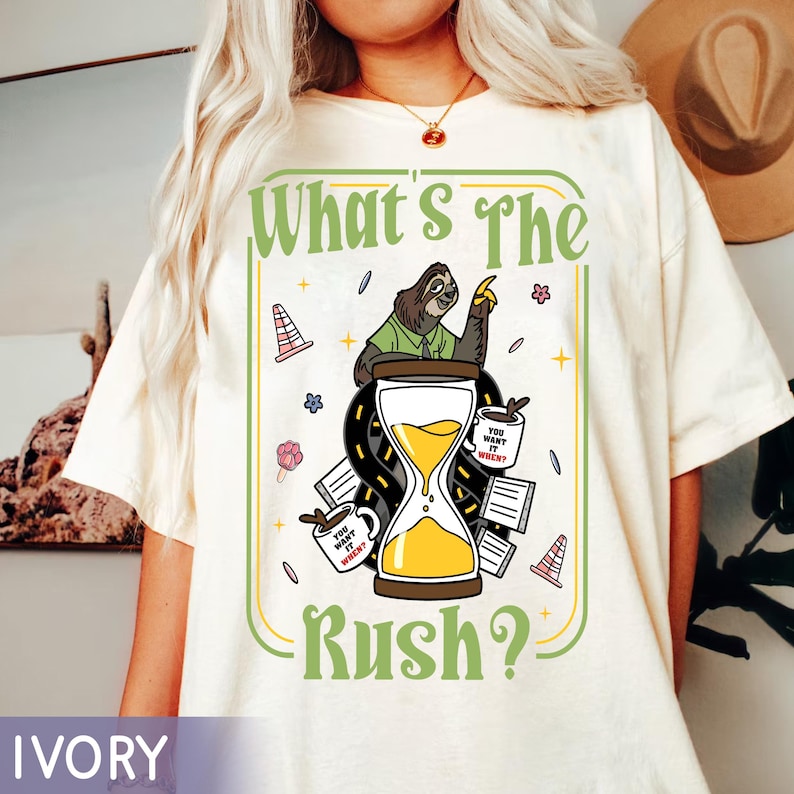 Puede incluir: Camiseta color marfil con un dise&ntilde;o gr&aacute;fico que presenta un perezoso se&ntilde;alando un reloj de arena. El texto "What's The Rush?" es verde, con gr&aacute;ficos adicionales de tazas de caf&eacute;, conos de tr&aacute;fico y flores.