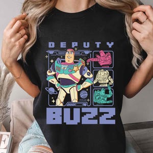 May include: Black t-shirt featuring a colorful graphic of Buzz Lightyear from the Pixar movie Toy Story. The design includes the words "DEPUTY" above and "BUZZ" below the character, along with other graphic elements.