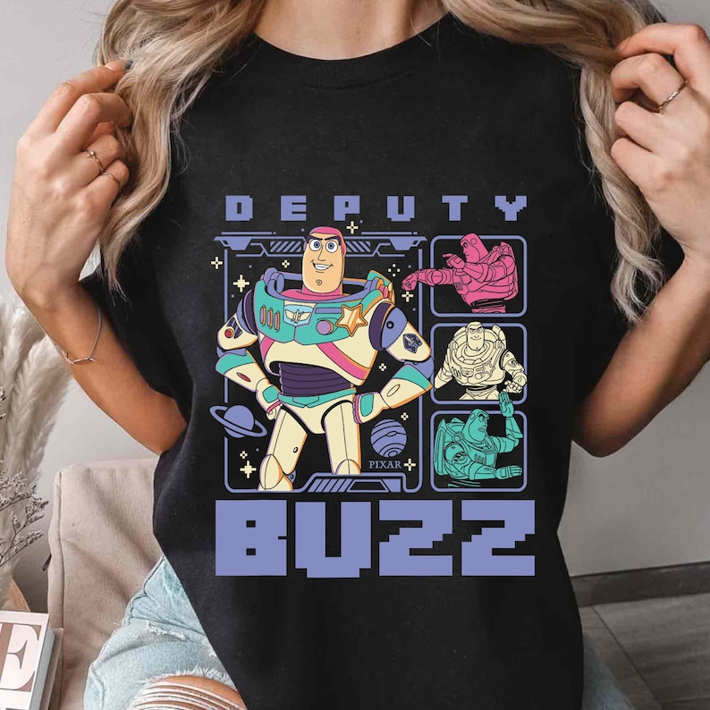 May include: Black t-shirt featuring a colorful graphic of Buzz Lightyear from the Pixar movie Toy Story. The design includes the words "DEPUTY" above and "BUZZ" below the character, along with other graphic elements.