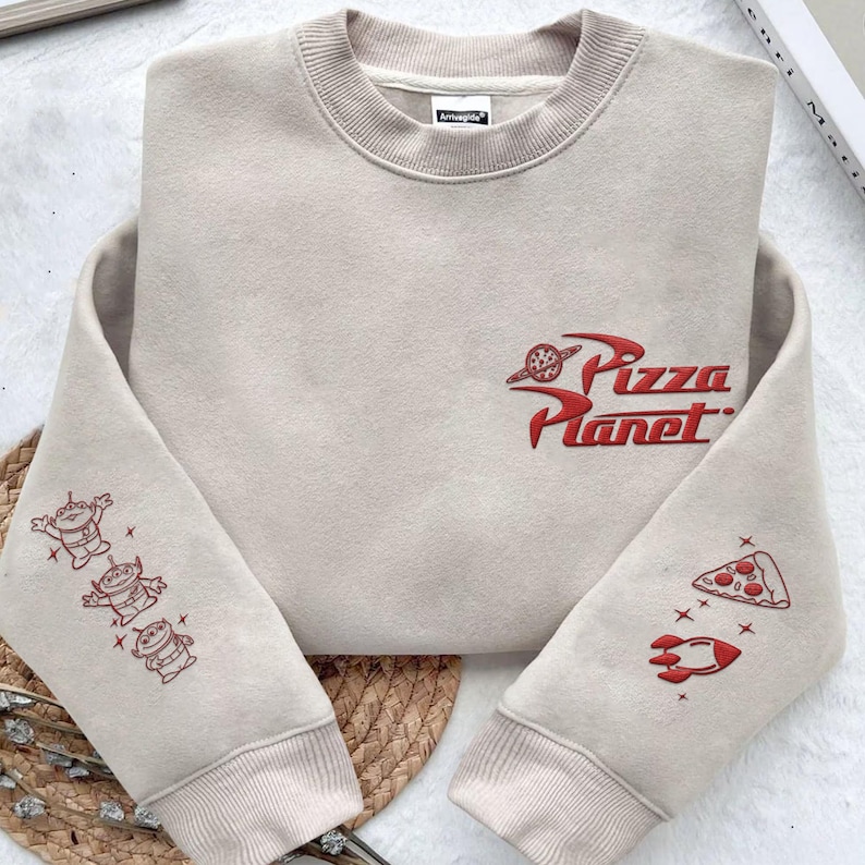 May include: A beige crewneck sweatshirt featuring red embroidered graphics. The chest displays the Pizza Planet logo. The left sleeve shows three small alien characters, while the right sleeve features a slice of pizza and a rocket ship design.