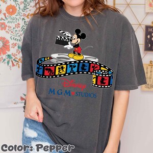 Pu&ograve; includere: T-shirt color pepe con Topolino che tiene una clapperboard, in piedi su una bobina di film. Una striscia di film colorata con personaggi Disney avvolge la bobina. Il logo Disney MGM Studios &egrave; sotto.