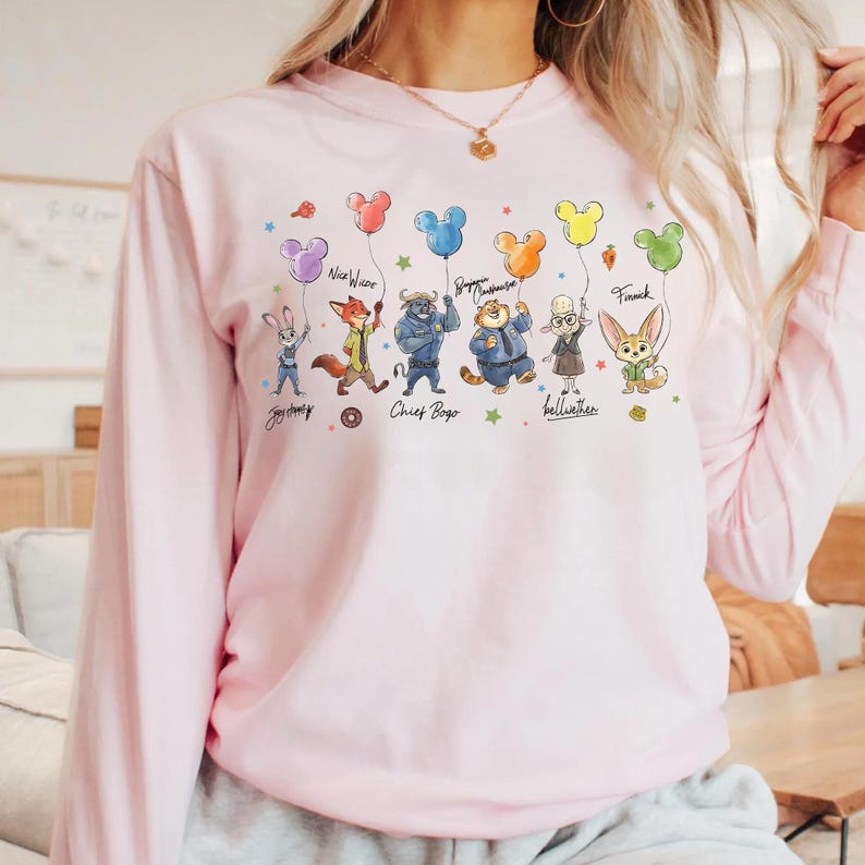 Op de afbeelding: Lichtroze sweatshirt met lange mouwen met een cartoonillustratie van personages die Mickey Mouse-vormige ballonnen vasthouden. Personages zijn onder meer Judy Hopps, Nick Wilde en Chief Bogo. De namen van de personages staan eronder.