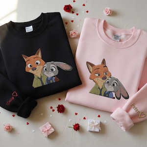 May include: Black and pink sweatshirts with embroidered cartoon fox and bunny characters. The black sweatshirt has "Daisy" embroidered on the sleeve, and the pink sweatshirt has "Peter" embroidered on the sleeve. Red roses and heart-shaped confetti are scattered around the sweatshirts.