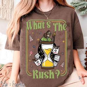Puede incluir: Camiseta color espresso con un dise&ntilde;o que presenta un perezoso, un reloj de arena y el texto "What's The Rush?". El dise&ntilde;o incluye tazas de caf&eacute; y elementos de carretera, creando un tema relajado. La camiseta es c&oacute;moda y casual.