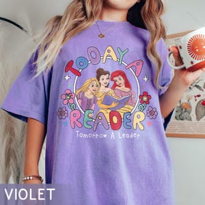 May include: Violet t-shirt with a graphic featuring three cartoon princesses reading a book, surrounded by flowers and the text "TODAY A READER Tomorrow A Leader."